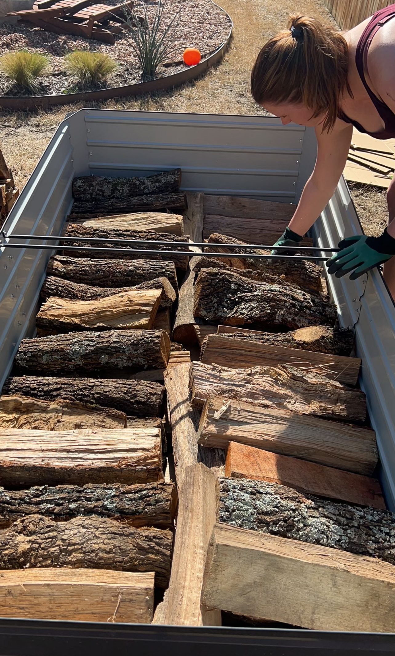 A person wearing gloves loads chopped firewood into a trailer. The trailer is filled with logs of various sizes and bark textures. The setting appears to be outdoors on a sunny day.