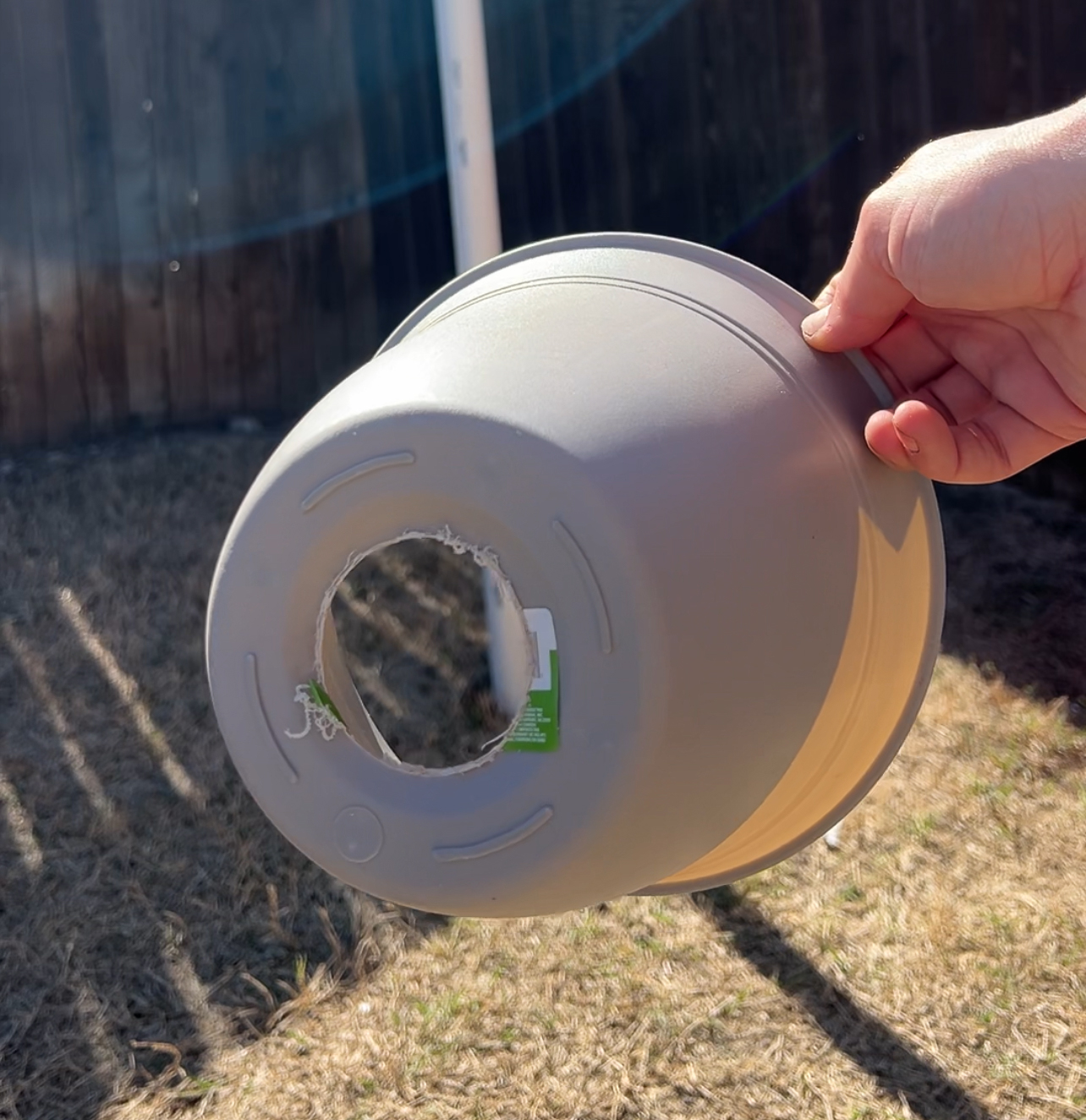 A hand holds a gray plastic bowl with a circular hole cut out of the base. The background features a sunlit grassy area and a wooden fence.