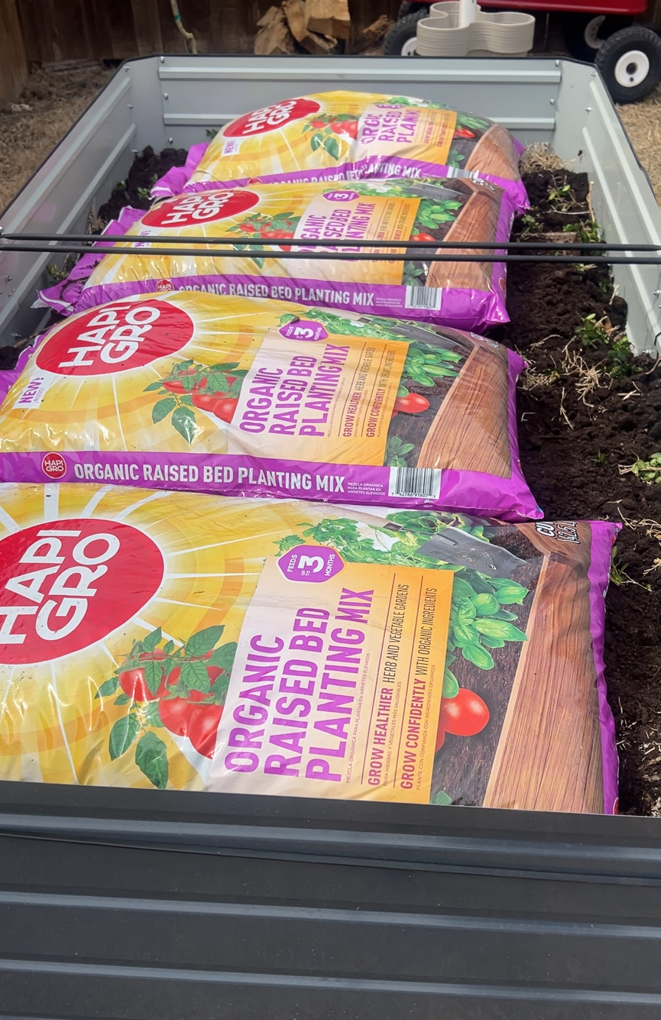 Bags of HAPI GRO Organic Raised Bed Planting Mix are laid out in a row on top of a garden bed filled with soil. The bags feature colorful labels with images of tomatoes and fresh produce.