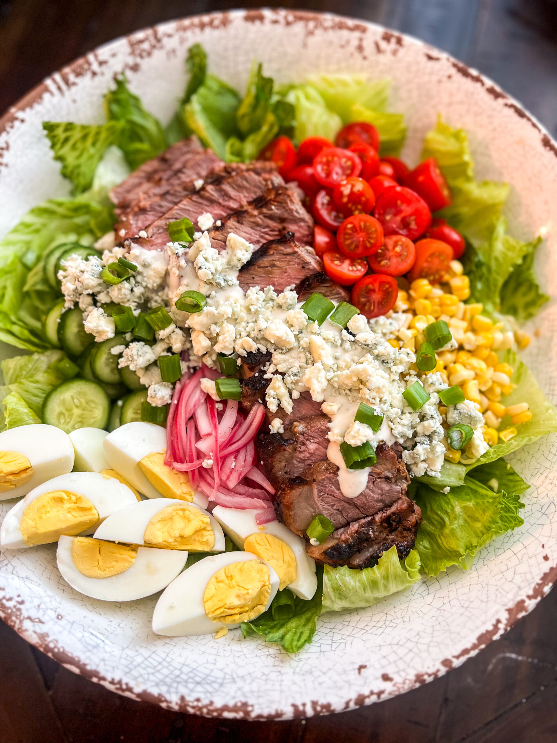 A vibrant black and blue steak salad with sliced steak, boiled eggs, pickled onions, cucumbers, cherry tomatoes, corn, blue cheese, and green onions atop crisp romaine lettuce in a round bowl.