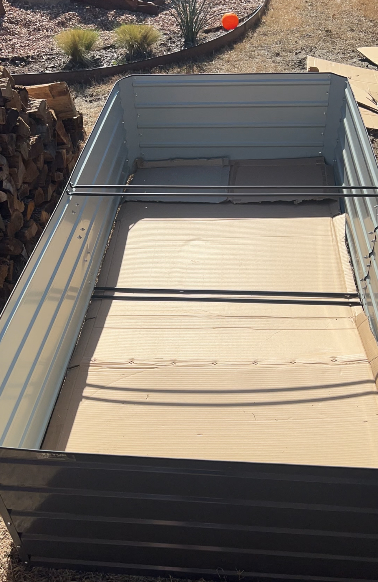 A metal framed raised garden bed under construction in a yard. The interior base is lined with cardboard. Firewood is stacked to the left, and the ground is dry with some vegetation in the background.