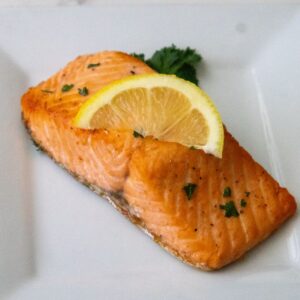 A piece of cooked salmon topped with a lemon slice and garnished with parsley is placed on a white plate.