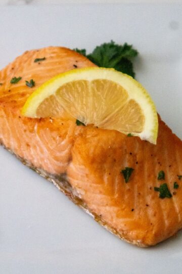 A piece of cooked salmon topped with a lemon slice and garnished with parsley is placed on a white plate.