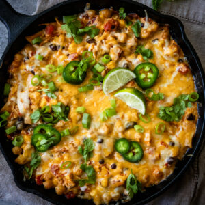 A skillet filled with a cheesy, baked dish topped with melted cheese, jalapeño slices, chopped green onions, cilantro, and lime wedges. Small corn kernels and other vegetables are visible, all baked to a golden brown.