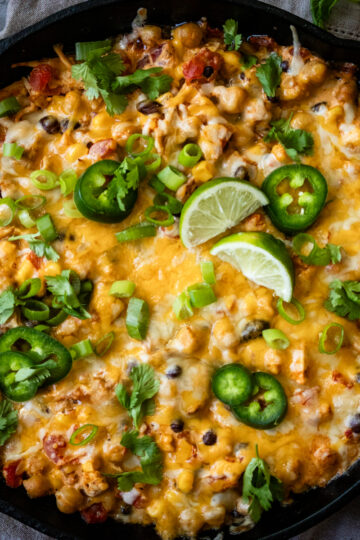 A skillet filled with a cheesy, baked dish topped with melted cheese, jalapeño slices, chopped green onions, cilantro, and lime wedges. Small corn kernels and other vegetables are visible, all baked to a golden brown.