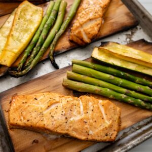 Grilled cedar plank salmon fillets served with asparagus spears and yellow squash slices, all seasoned and roasted to perfection on a metal baking tray.