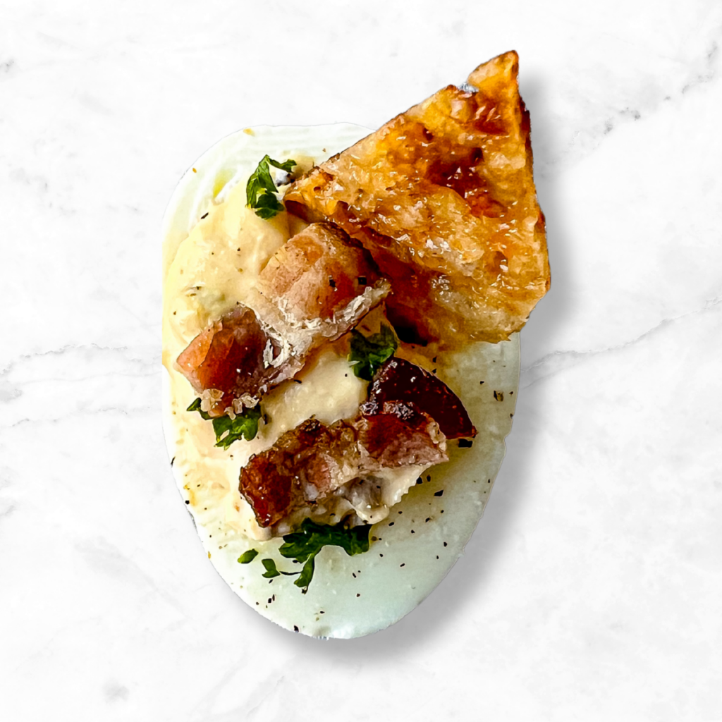 A deviled egg topped with crispy bacon pieces, fresh herbs, and a crunchy triangular chip, set on a white marble background.