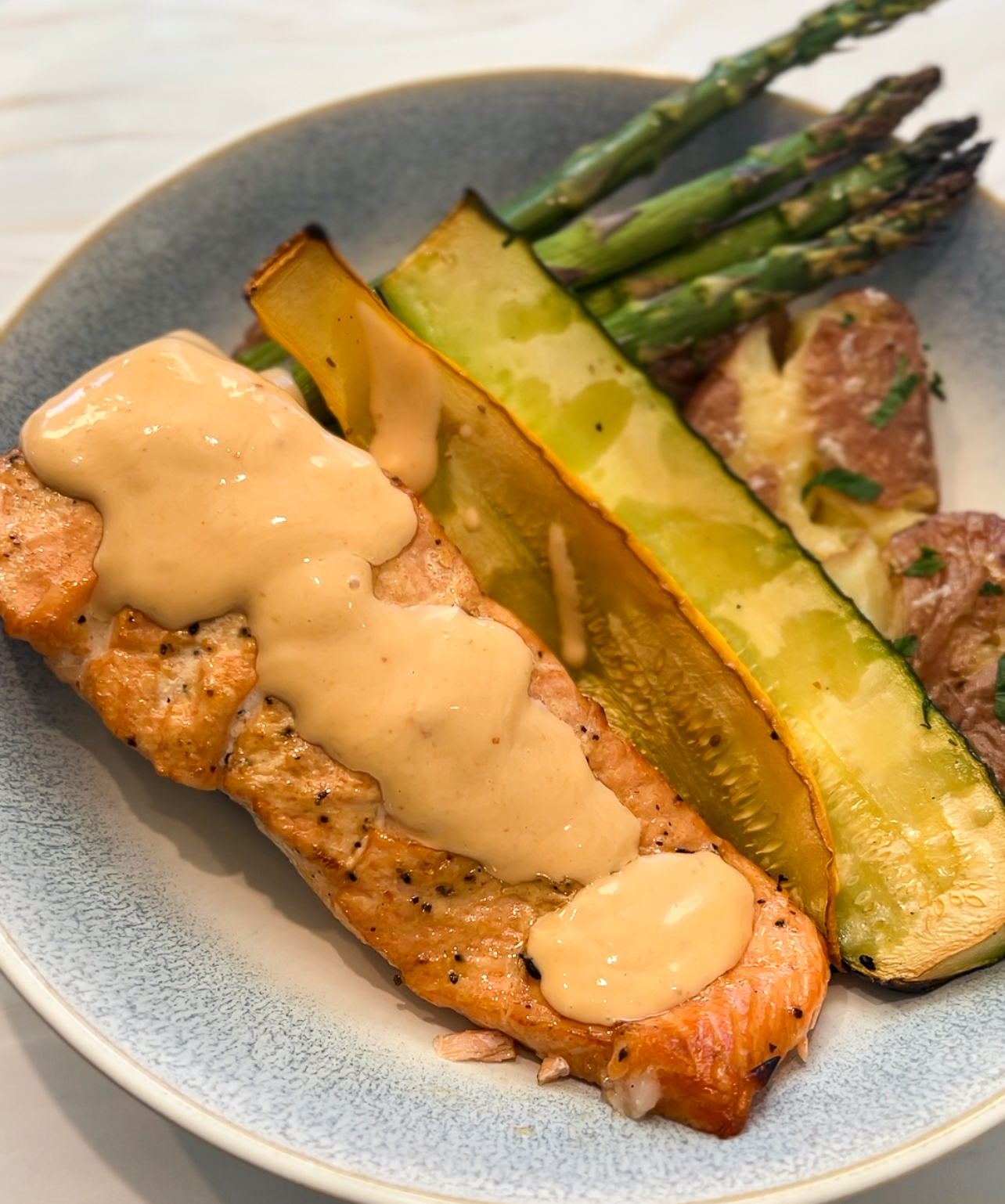 A plate with grilled bang bang salmon topped with creamy sauce, accompanied by roasted zucchini, asparagus, and roasted potatoes.