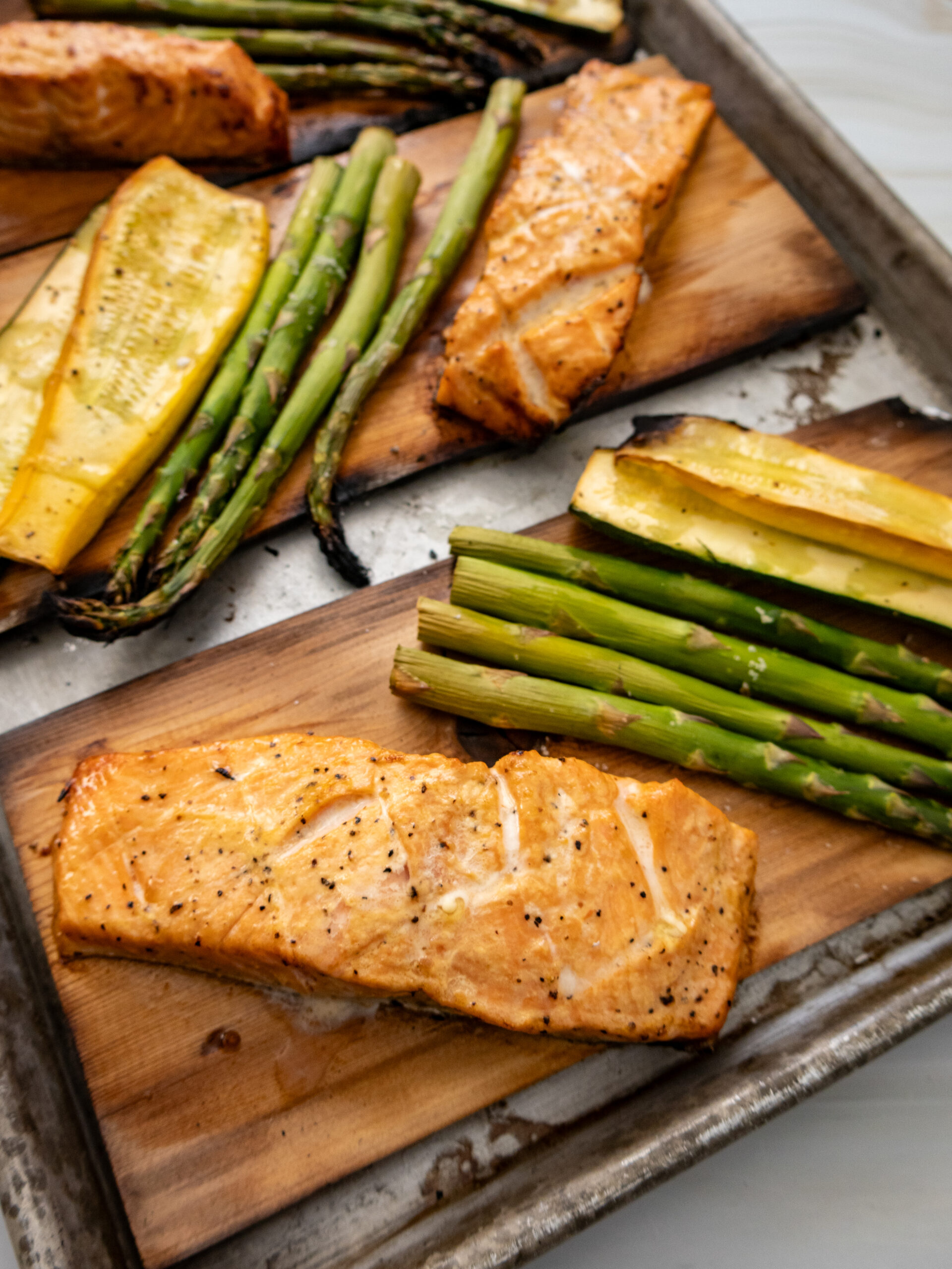 Grilled bang bang salmon fillets served on wooden planks with asparagus spears and yellow squash slices on a metal baking tray.