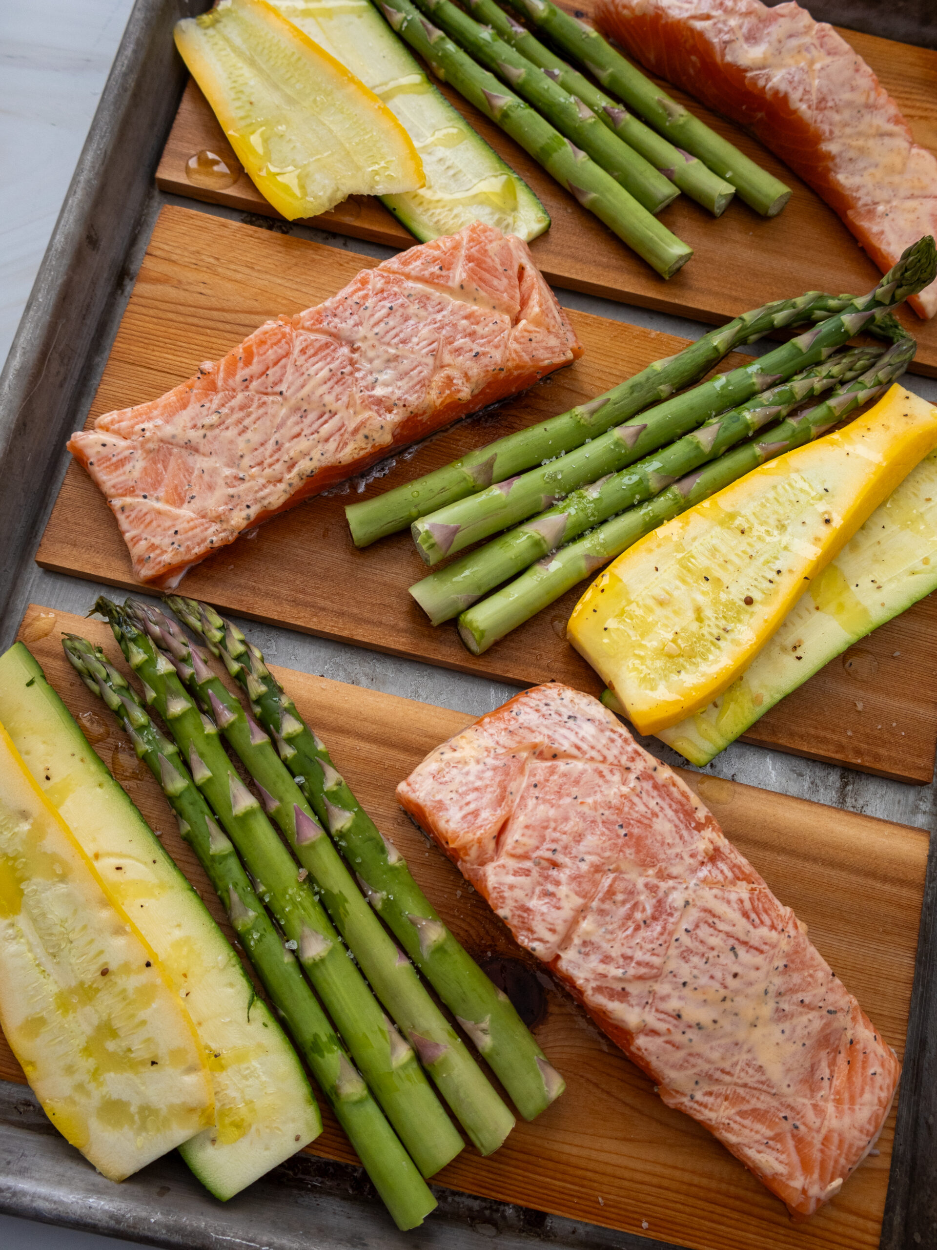 Raw salmon fillets and asparagus spears are arranged on a baking sheet with wooden planks for classic cedar plank salmon; yellow squash slices join the medley, all lightly seasoned with oil and pepper, ready to be cooked to perfection.