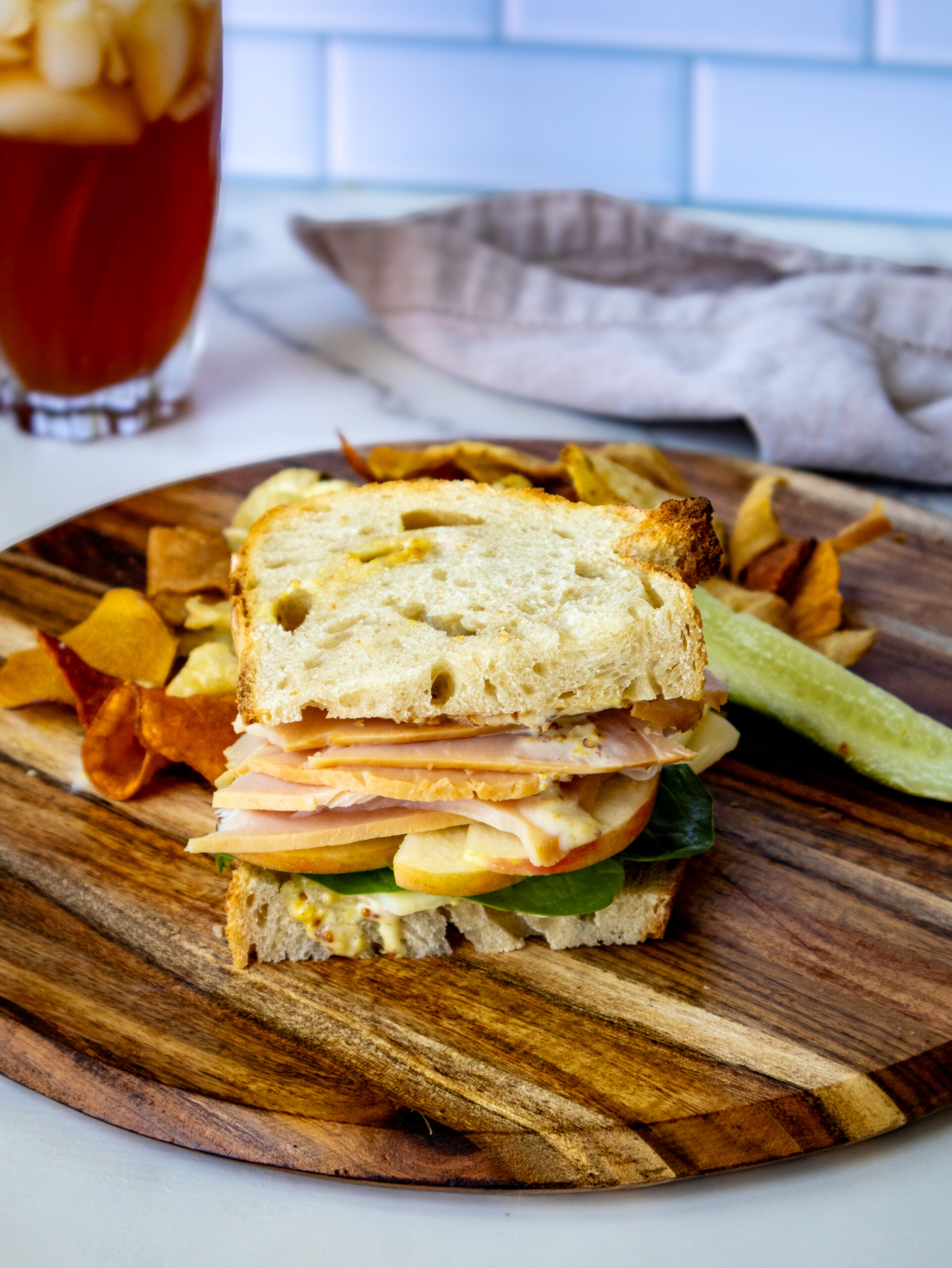 A turkey apple cheddar sandwich with lettuce on rustic bread sits on a wooden board, accompanied by potato chips and a pickle spear. A glass of iced tea and a napkin are in the background.
