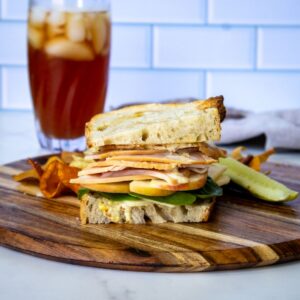 A turkey apple cheddar sandwich with lettuce and tomato on rustic bread sits on a wooden board with chips and a pickle spear, alongside a glass of iced tea in the background.