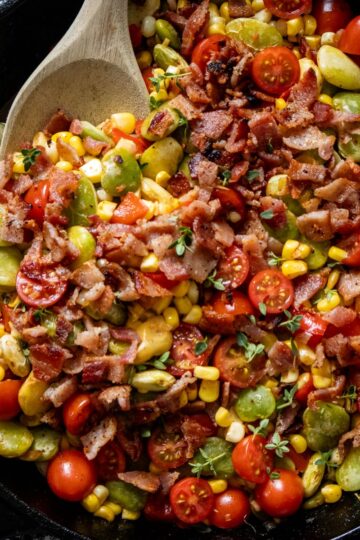 A skillet filled with a colorful corn succotash—a mix of corn, cherry tomatoes, lima beans, and crispy bacon pieces—makes the perfect summer succotash side dish, with a wooden spoon resting inside.