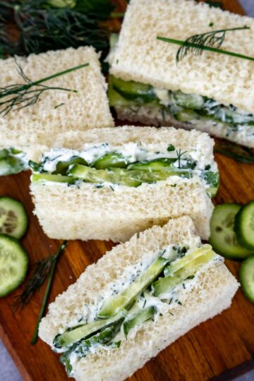 Classic cucumber sandwiches with herb cream cheese are arranged on a wooden board, surrounded by fresh cucumber slices and sprigs of dill and chives.