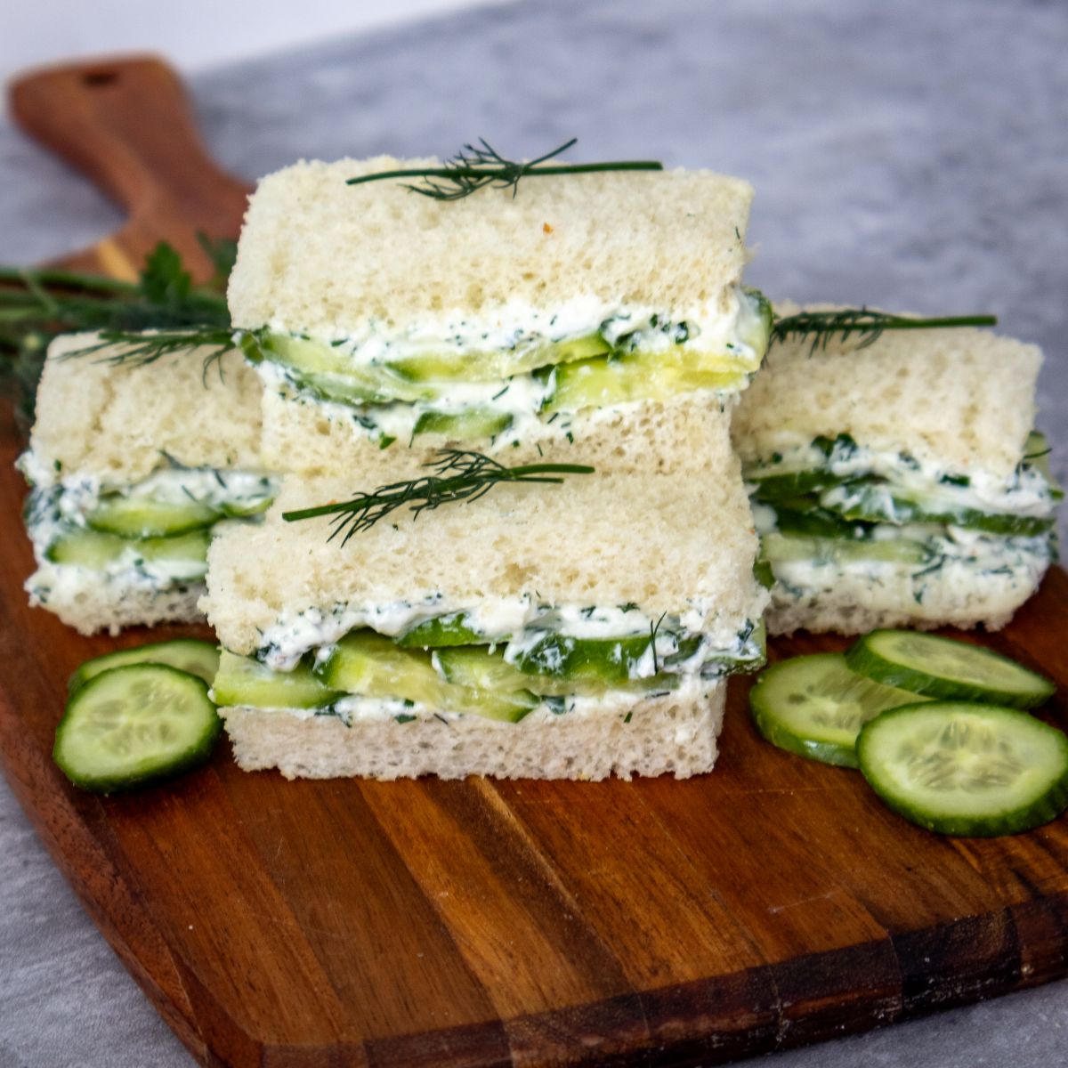 Three classic cucumber sandwiches with herb cream cheese are stacked on a wooden board, garnished with fresh dill and cucumber slices. The bread is white and crustless, and the sandwiches are neatly cut into rectangles.