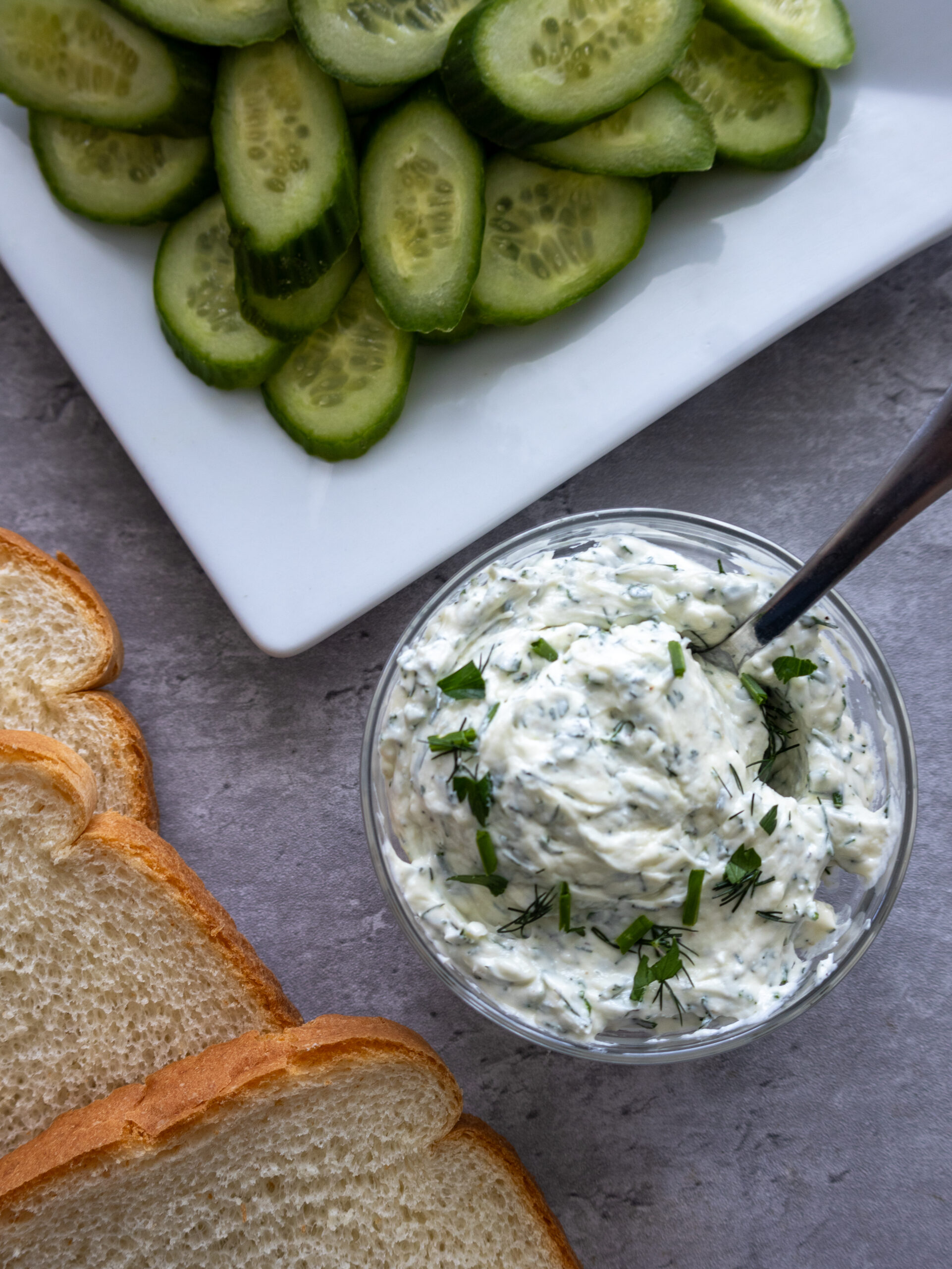 A bowl of creamy herb spread with a spoon, garnished with fresh herbs, sits next to slices of white bread and a plate of cucumber slices—perfect for making a classic cucumber sandwich or a delightful cucumber cream cheese sandwich.