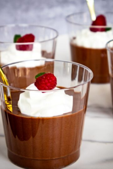 Five clear cups filled with chocolate mousse, topped with whipped cream and a raspberry, each with a gold spoon, arranged on a white surface.