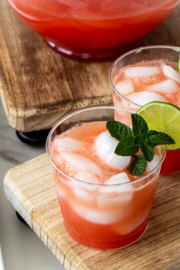 Two glasses of iced watermelon punch garnished with lime slices and mint leaves sit on a wooden board, with a punch bowl and watermelon slices nearby.